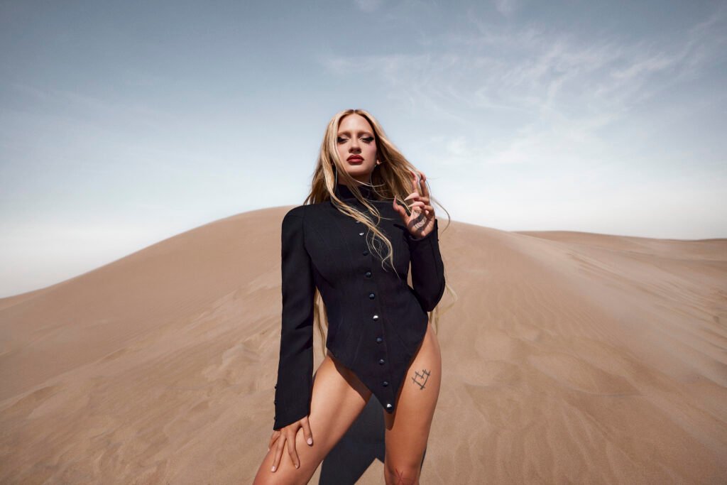 La artista Luna Ki posando en medio de un desierto con dunas de arena, vistiendo un body negro de manga larga.