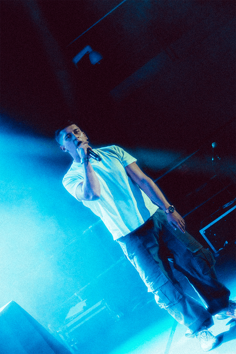 El artista Nakku cantando con un micrófono, iluminado por una potente luz de escenario azul o cian durante una actuación.
