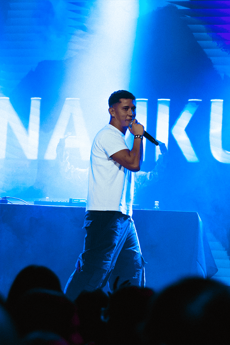 El artista Nakku cantando sobre el escenario, con su nombre proyectado en las pantallas de fondo durante un concierto.