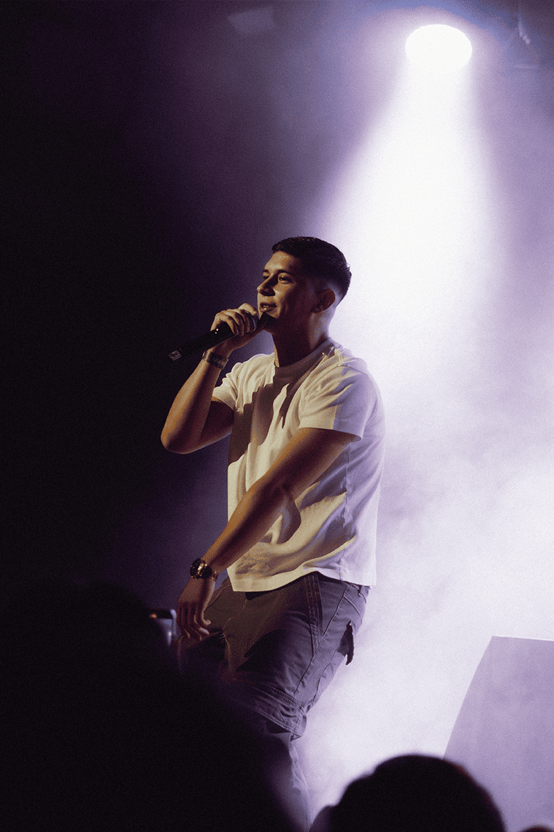 El artista Nakku cantando en directo bajo un foco de luz cenital, con una camiseta blanca sobre el escenario.