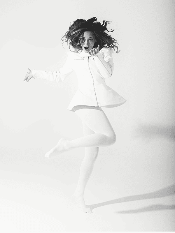 Fotografía en blanco y negro de la artista Luna Ki saltando en el aire con un conjunto blanco, en una pose enérgica.