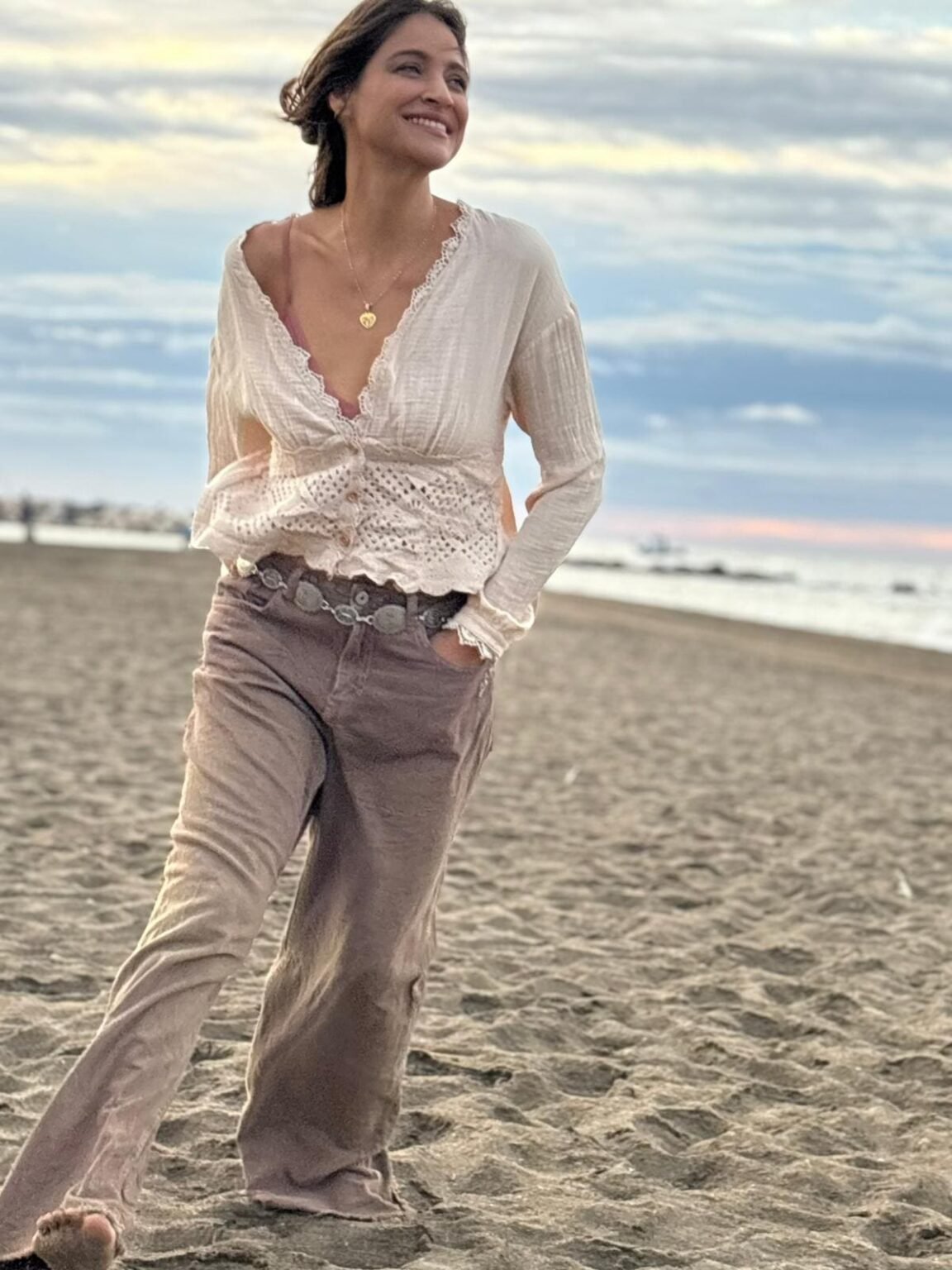 La cantante Georgina sonriendo en una playa durante el atardecer, vestida con una blusa blanca y pantalones anchos.